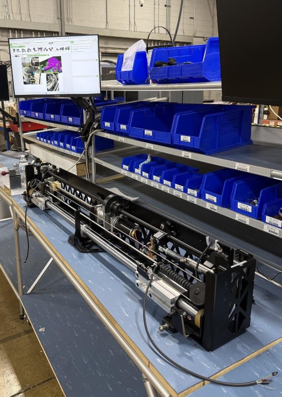 The Gen 4 product on an assembly bench next to a bin rack and a screen showing the work instructions